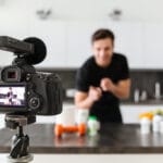 Happy young man filming his video blog episode about healthy food additives while standing at the kitchen table and pointing finger at camera